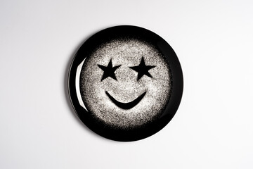 Smiling face, concept made with plate and flour, white background, star shaped eyes, black plate, happy mood