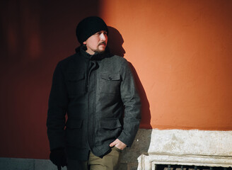 A young guy in a gray coat and hat leans on a terracotta-colored wall in the rays of the setting...