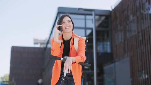 Emotional caucasian woman dressed in casual bright attire talking on modern smartphone while leaning on handle of electic scooter on fresh air. Concept of technology and youth lifestyles.