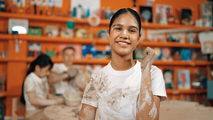Happy caucasian girl pose at camera while diverse children modeling clay behind. Cute student wearing dirty shirt while looking at camera at workshop in art lesson. Blurring background. Edification. - Powered by Adobe