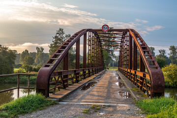 A steel bridge over a water channel connecting dirt roads. There is a sign on the bridge specifying the maximum weight limit for a passing vehicle.