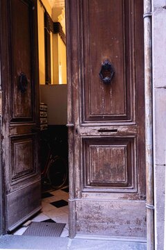 Portal con puerta antigua de madera entreabierta, con aldaba, al fondo se ven unos buzones y el suelo ajedrezado, vertical
