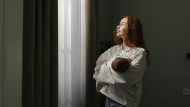 Mom Rocking Baby In Arms. Woman Enjoys View From Window, Rocks Baby In Arms, Helping To Fall Asleep. Young Happy Mom Feels Good Doing Mom Duties. Parenthood, Motherhood, Parenting And Childcare.