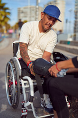 athlete man with disability in wheelchair putting on knee protection with help at skatepark	