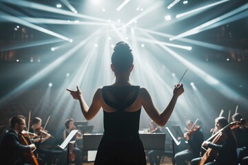 Female conductor leads an orchestra, baton in hand, under dramatic stage lights in a grand concert or philharmonic hall - obrazy, fototapety, plakaty