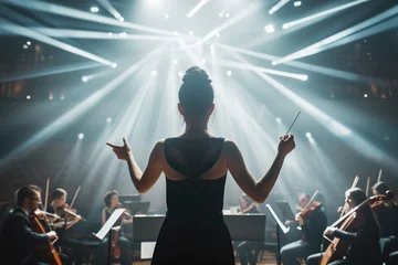 Fotobehang Female conductor leads an orchestra, baton in hand, under dramatic stage lights in a grand concert or philharmonic hall © iridescentstreet