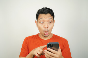 Wow face of Asian man in orange t-shirt shocked what he see in the smartphone on isolated background