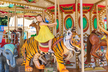 Fototapeta na wymiar Adorable little blonde girl in summer yellow dress at amusement park having a ride on the merry-go-round. Child girl has fun outdoor on sunny summer day. Entertainment concept
