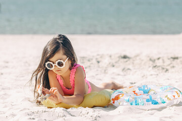 Happy cute smiling little girl in sunglasses lying on the sand on a beach in a swimsuit. Summer...