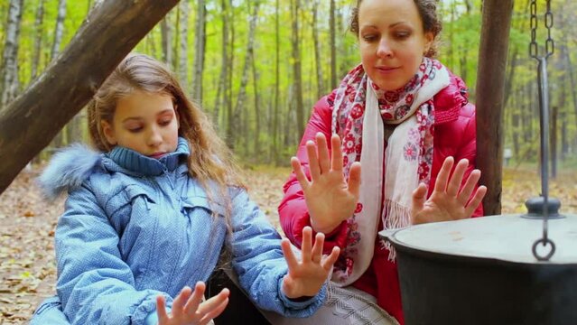 Woman and girl warm hands near kettle and smolder log