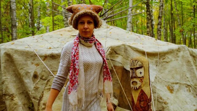 Woman in native fur hat looks on idol near wigwam at autumn day in forest