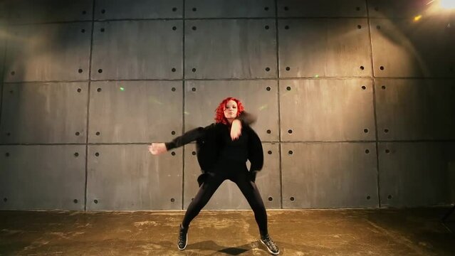 Red-haired girl in black clothes dances near wall in studio