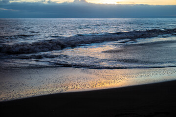 Obraz premium A sunset on Waimea Beach on Kauai reflects warmth onto gentle waves and a glow on the dark sand.