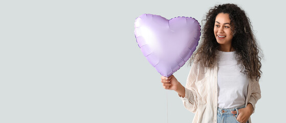 Beautiful young African-American woman holding heart-shaped balloon on light background with space...