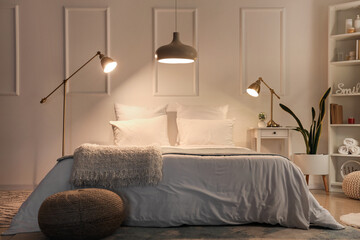 Interior of cozy bedroom with soft blankets on comfortable bed, wicker pouf and glowing lamps at...