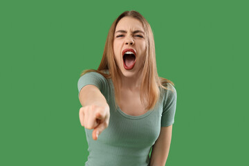 Angry young woman pointing at viewer on green background. Accusation concept - obrazy, fototapety, plakaty