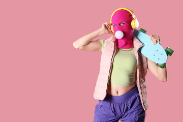 Beautiful young stylish woman in balaclava with headphones and skateboard chewing gum on pink...