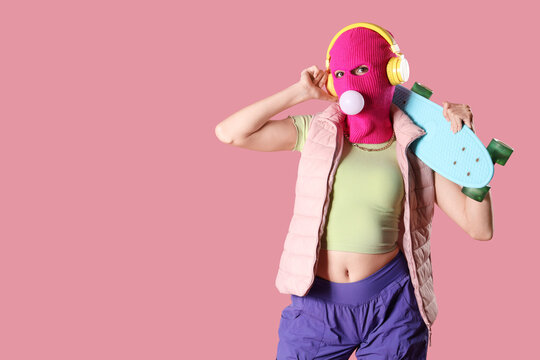 Beautiful young stylish woman in balaclava with headphones and skateboard chewing gum on pink background