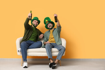Young men in leprechaun hats with green beards holding bottles of beer and sitting on sofa near...