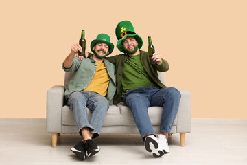 Happy young men in leprechaun hats with green beards holding bottles of beer and sitting on sofa...