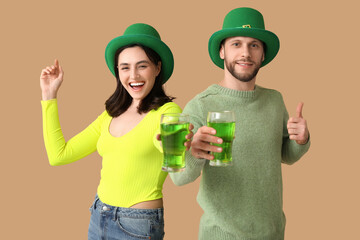 Beautiful young couple in leprechaun hats with glasses of beer showing thumb-up gesture on brown...