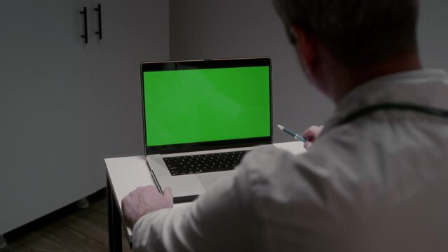 Professional male doctor in work uniform looks at laptop screen listening to patient and pointing with pen to image area. Chromakey for inserting photo and video concept slow motion