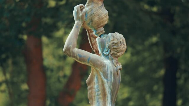 A statue of a woman drinking from the amphora on the blurry background. The statue is covered with patina.