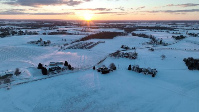 Winter snow covers rural country fields in Northeast USA. Starburst sunrise with farming chicken houses and country road. Aerial view.