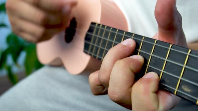 Man mid 30 and with bead is playing and whistling with his pink ukulele next to a big window and close to a green plant. Music is one form of art that can soothe the soul and make us feel better