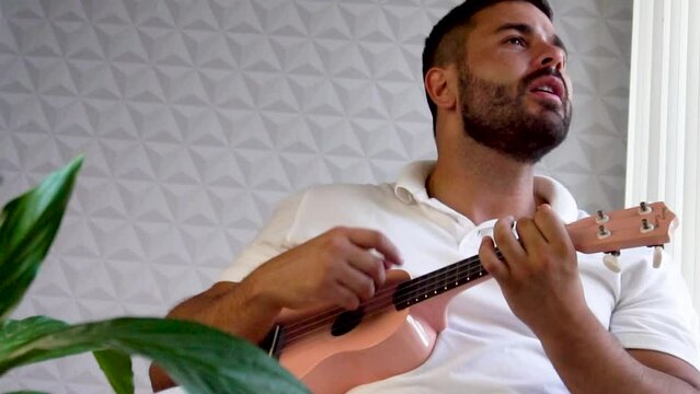 Man mid 30 and with beard is playing and singing with his pink ukulele next to a big window and close to a green plant. Music is one form of art that can soothe the soul and make us feel better