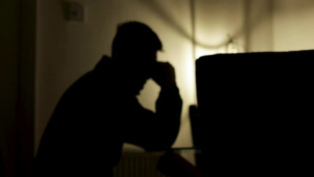 Silhouette of distressed young man sat at table with atmospheric lighting, with his head in hands. Static.