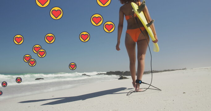 Image of heart digital icons over woman carrying surfboard on beach