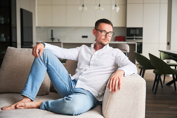 Charming man relaxing on couch in his apartment. Handsome young male thoughtfully looking away. Man...