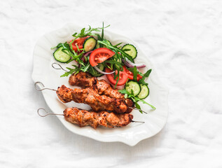 Delicious lunch, appetizer, tapas - chicken kebabs and fresh vegetable cucumbers tomatoes salad on a light background, top view