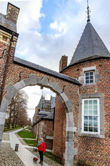 Exterior path of ancient 16th century Alden Biesen castle, female tourist standing with her dog under entrance arch, bare trees in background, cloudy day in Bilzen, Limburg, Belgium