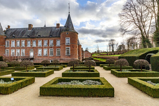 French garden with bushes and small trees, 16th century Alden Biesen Castle in background, bare trees, windows, brick walls, gable roof and circular towers, cloudy day in Bilzen, Limburg, Belgium