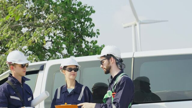 Group of technician workers stand beside the van and discuss about work with windmill or wind turbine on the background.