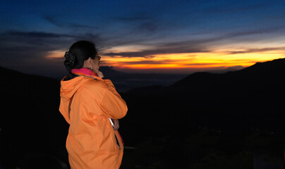 Female mountain climber in orange jacket standing alone to welcome the morning sun reflected on...