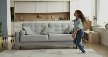 Excited active dancer kid girl hopping past camera in modern comfortable apartment, dancing at couch, moving through living room with spacious kitchen in background, enjoying new home