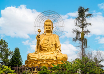 golden Buddha sculpture stands tall with lotus in hand, against backdrop of stunning sky and surrounded by lush tropical plants. radiant sculpture exudes tranquility and spirituality