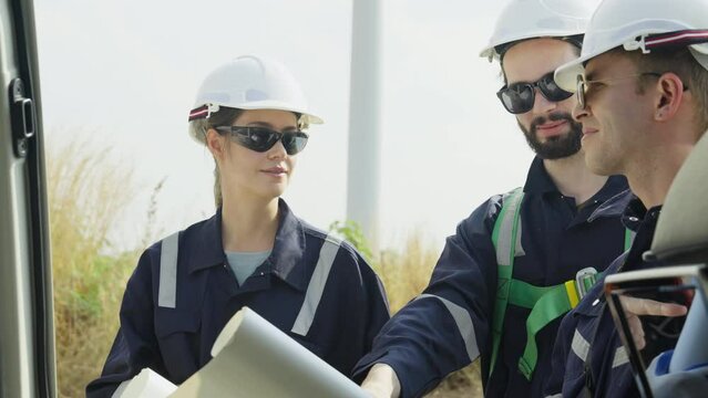 Close up group of technician workers stand beside the van that open the door and discuss about work using plan paper with windmill or wind turbine on the background.