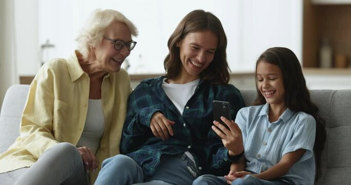 Happy grandma, mother and cheerful girl holding smartphone and laughing out loud, looking at screen, having fun, relaxing on comfortable sofa, using funny media service, online application for selfie
