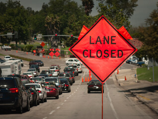 An orange diamond shaped roadwork sign statiing lane closed with traffic backed up in the distance