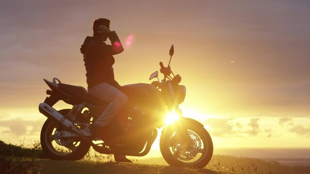 Man sitting on motorbike removes helmet and looks at sunset
