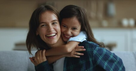 Joyful cute girl embracing happy young mom from behind, looking at camera with toothy smile. Cheerful mother piggybacking kid, enjoying motherhood, playtime, leisure with beloved child