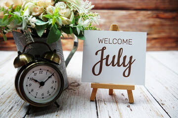 Welcome July text message written on paper card with wooden easel and alarm clock with flower in metal vase decoration