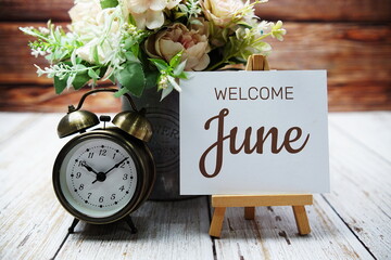 Welcome June text message written on paper card with wooden easel and alarm clock with flower in metal vase decoration - obrazy, fototapety, plakaty
