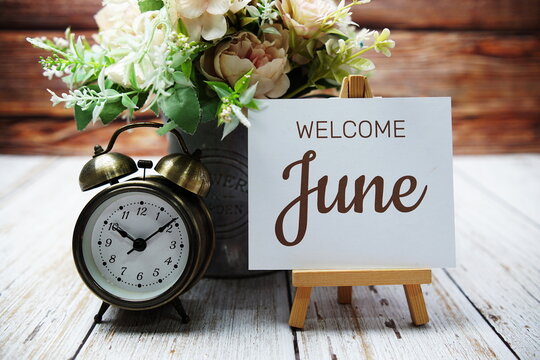 Welcome June text message written on paper card with wooden easel and alarm clock with flower in metal vase decoration