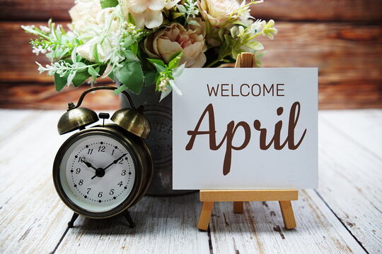 Welcome April text message written on paper card with wooden easel and alarm clock with flower in metal vase decoration