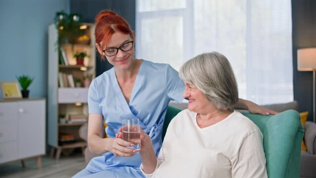 portrait of young female social worker brings a glass of water to an elderly woman, smiling and looking at camera